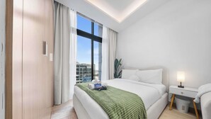A modern hotel room with a large bed, a bedside table with a lamp, and a view of the city through the window. - Stayshort - Studio Apartment at Azizi Riviera With Balcony (Dubai)
