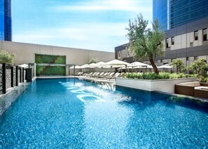 A swimming pool with lounge chairs and umbrellas, surrounded by modern buildings. - Stayshort - Chic Studio for 3 People in Business Bay (Dubai)
