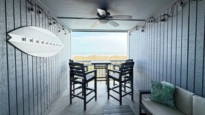 Un espace extérieur abrité, doté de sièges, offrant une vue sur la plage, d’un ventilateur de plafond et d’une décoration sur le thème du surf.