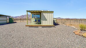 A small, single-room structure with a window and a door, a small porch with two chairs, and a gravel ground. - Tiny Town Cottages in Rodeo! 'Dog House' (Rodeo)