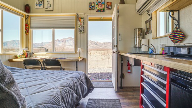 A bedroom with a large bed, a desk, and a view of the desert through an open door. - Tiny Town Cottages in Rodeo! 'Dog House' (Rodeo)
