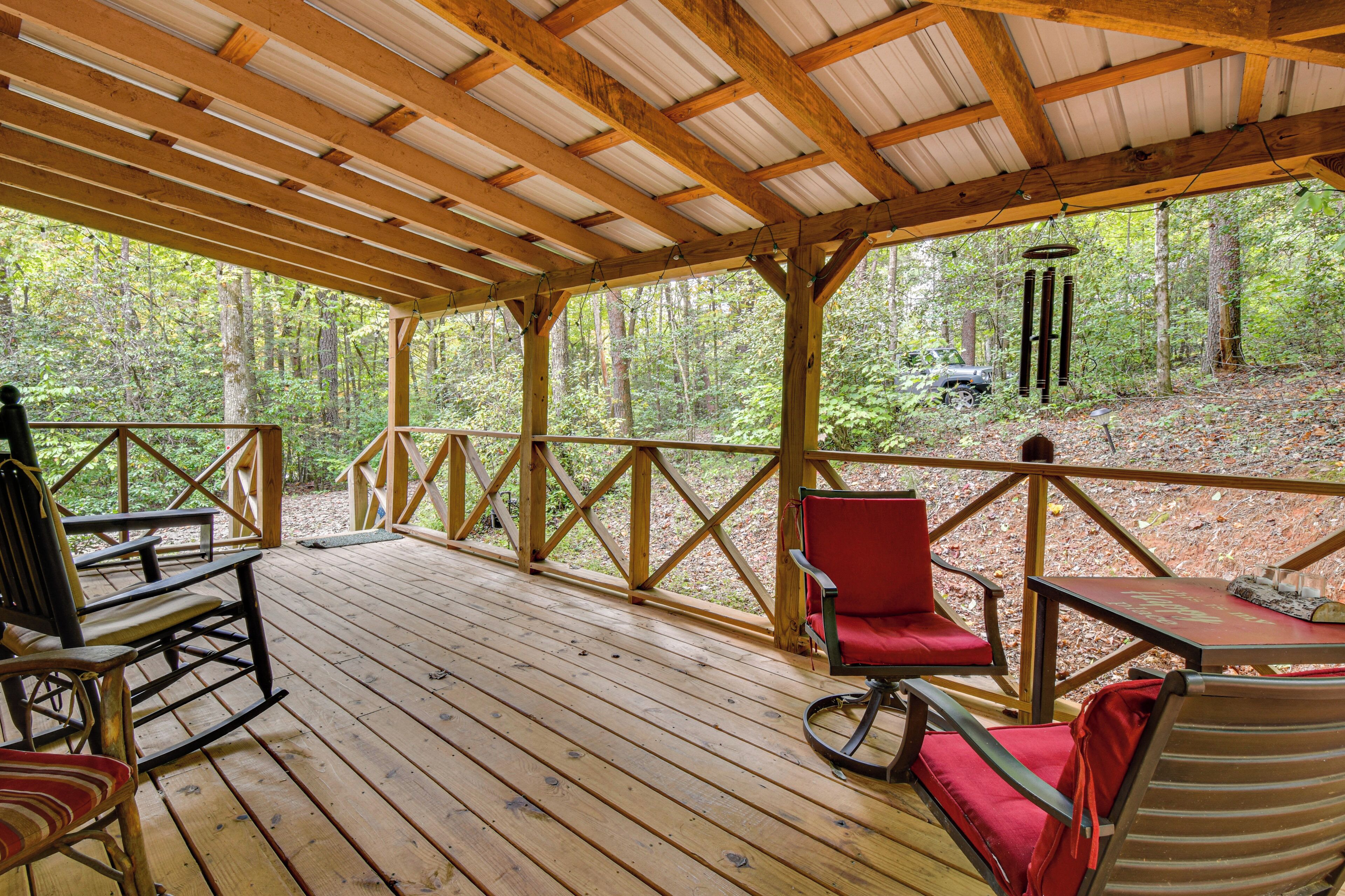 Terraza de madera con un área cubierta, amueblada con mecedoras y una mesa, con vista a un bosque.