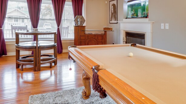 A pool table in a room with wooden floors, a fireplace, and a window with purple curtains. - Centrally Located Hampton Roads Home w/ Game Room! (Portsmouth)