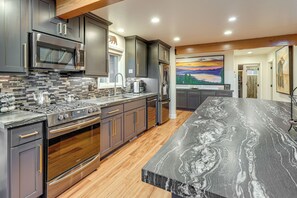 A modern kitchen with dark cabinetry, stainless steel appliances, a large marble countertop, and a painting of a sunset on the wall. - Retreat w/ Hot Tub: Bear Mountain Ski Slope View (Big Bear Lake)