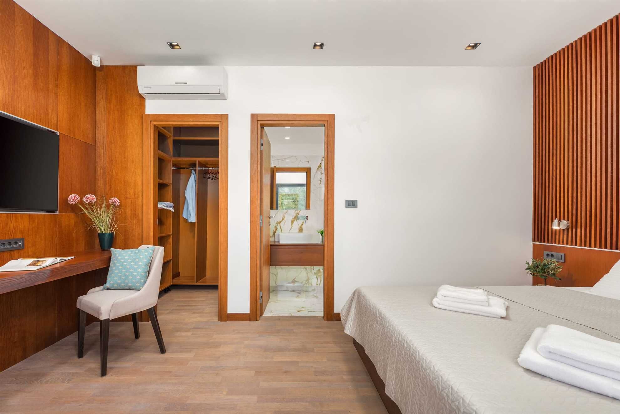 A hotel room with a wooden wardrobe, a bed with white linens, a desk with a chair, and a TV mounted on the wall.