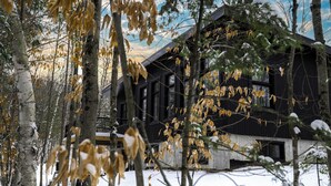 Une cabane rustique aux murs en bois, entourée d’un paysage enneigé et d’arbres aux feuilles d’automne.