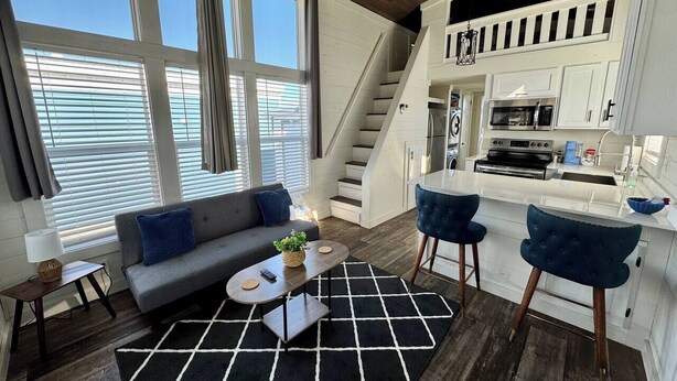 Un salon moderne avec un canapé, une table basse et deux fauteuils. Un escalier mène à un espace cuisine équipé d’un four à micro-ondes et d’une cuisinière.