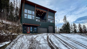Un edificio moderno de dos pisos con un gran portón de garaje, rodeado de nieve y altos árboles.
