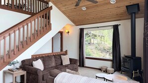 A cozy living room with a wooden staircase, a ceiling fan, a large window with a view, and a wood-burning stove. - Unwind at Lakeside Hideaway overlooking CDA Lake (Harrison)