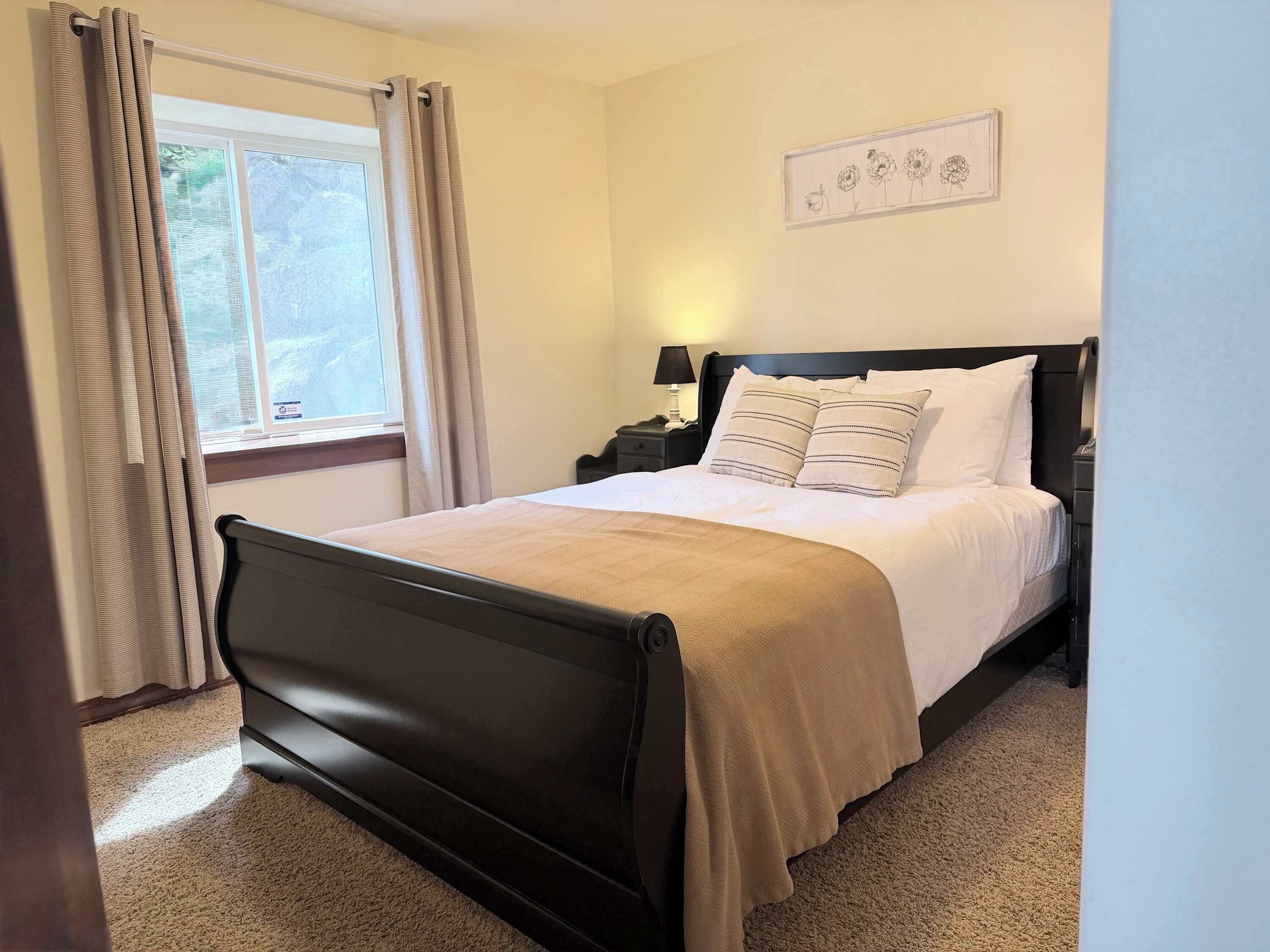 Un dormitorio con cama, mesita de noche, lámpara, ventana con cortinas e imagen colgada en la pared.