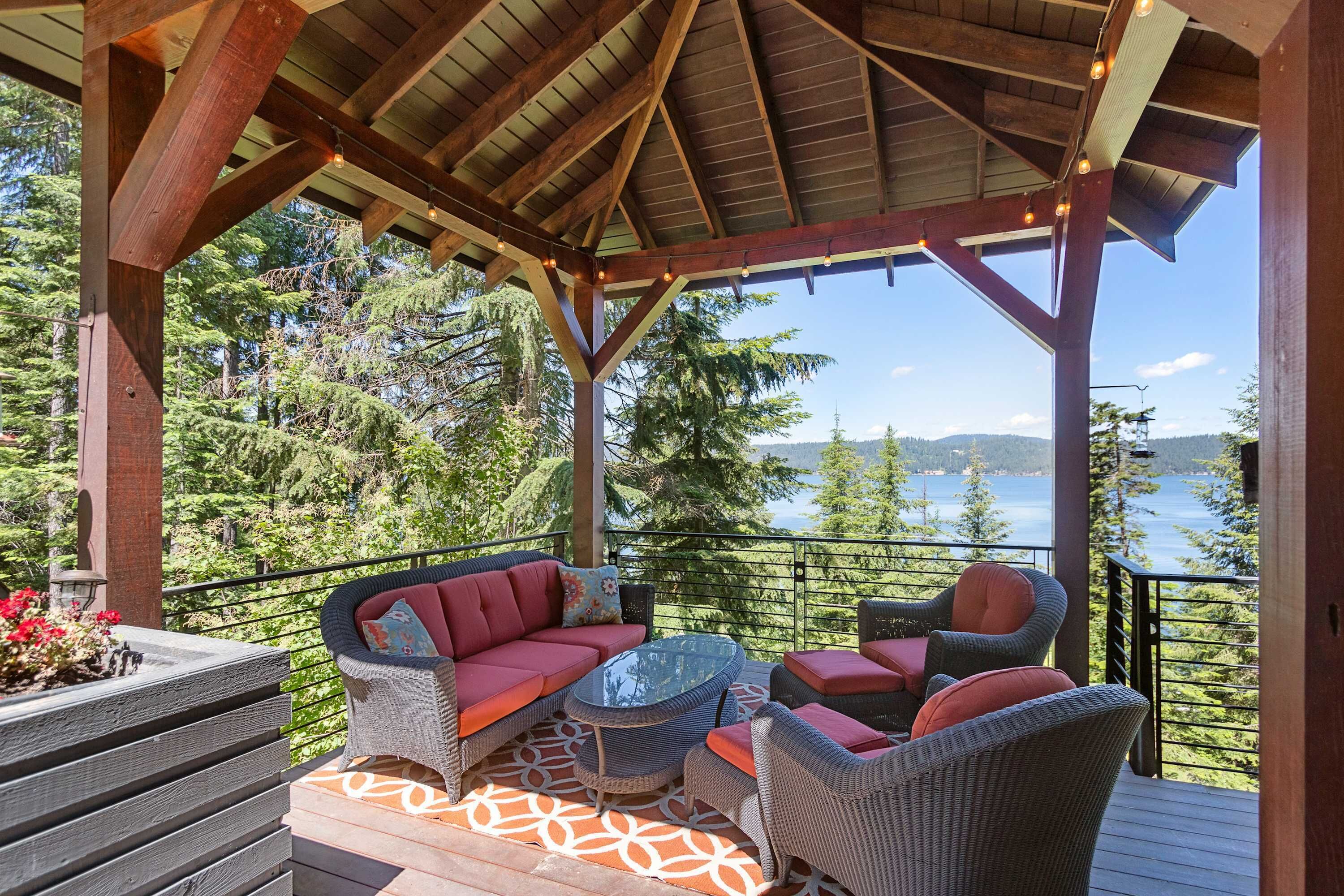 A covered deck with wicker furniture, a glass table, and a view of a lake and forest.