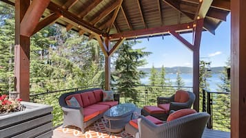 A covered deck with wicker furniture, a glass table, and a view of a lake and forest.