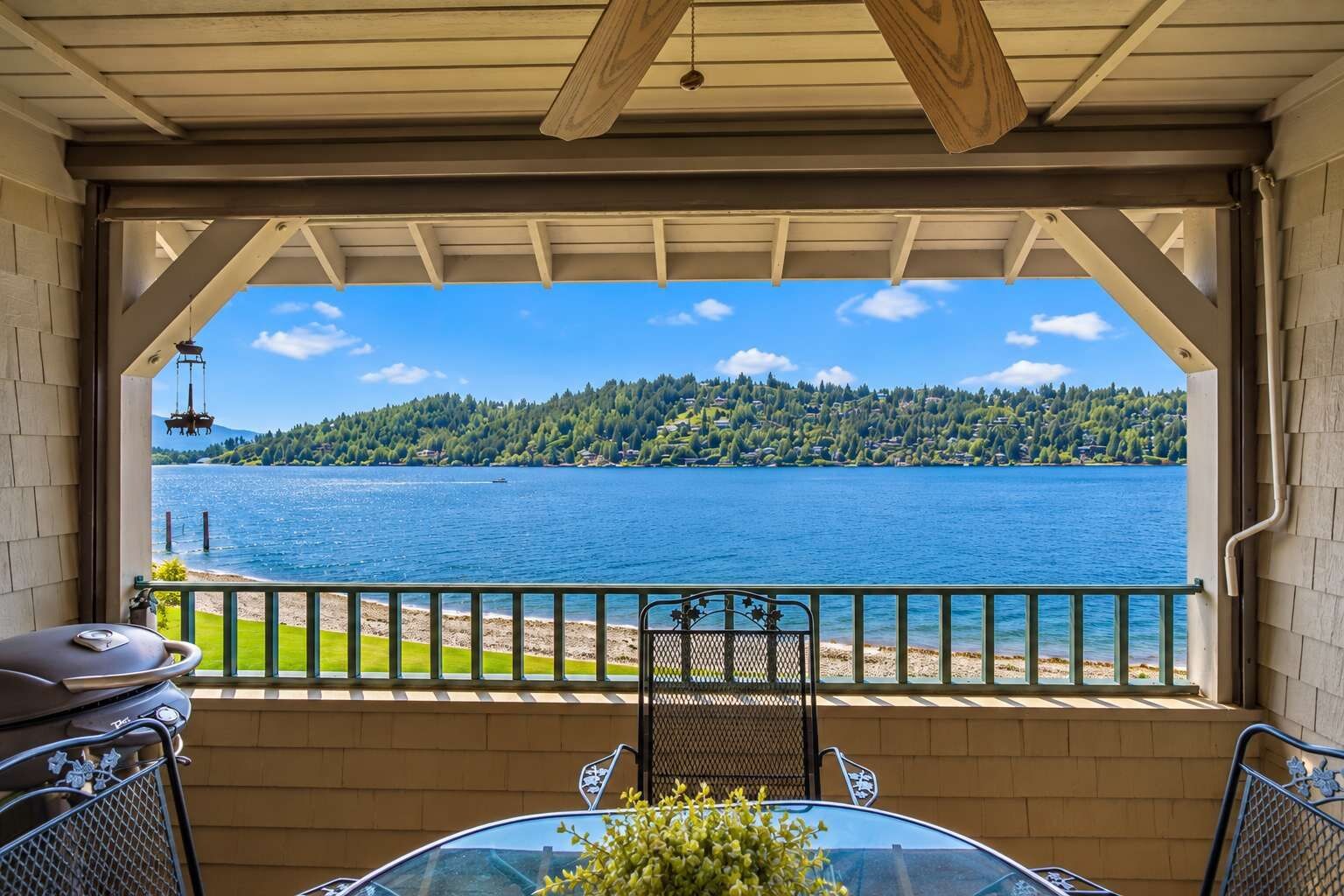 A covered outdoor area with a view of a lake, a grill, and a table with chairs.