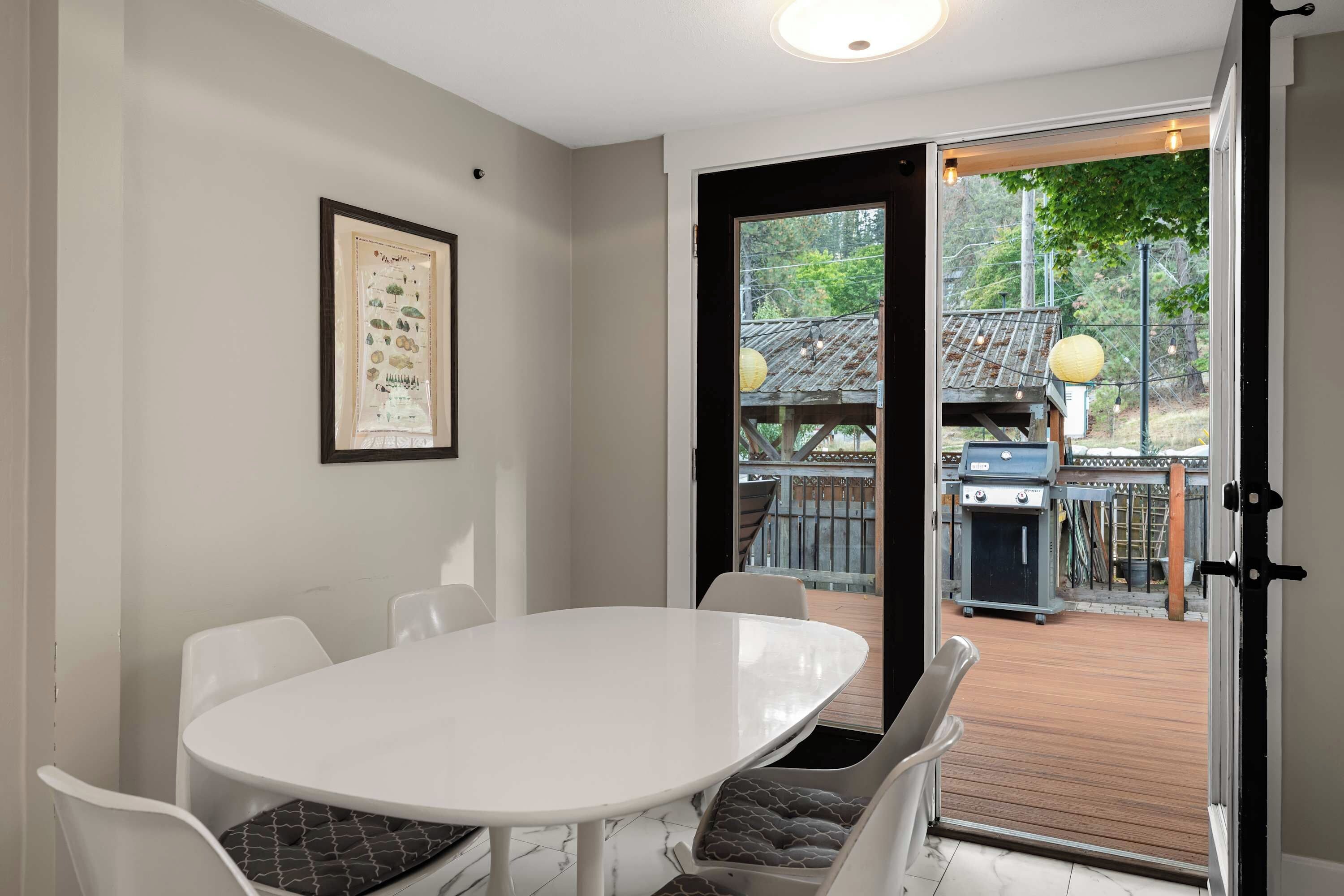 A dining area with a white table and chairs, a framed poster on the wall, and a view of an outdoor deck with a grill.