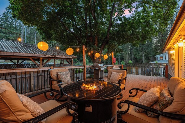 Un patio exterior con una mesa redonda y sillas, un hogar para fogata, luces colgantes y vistas de una casa.