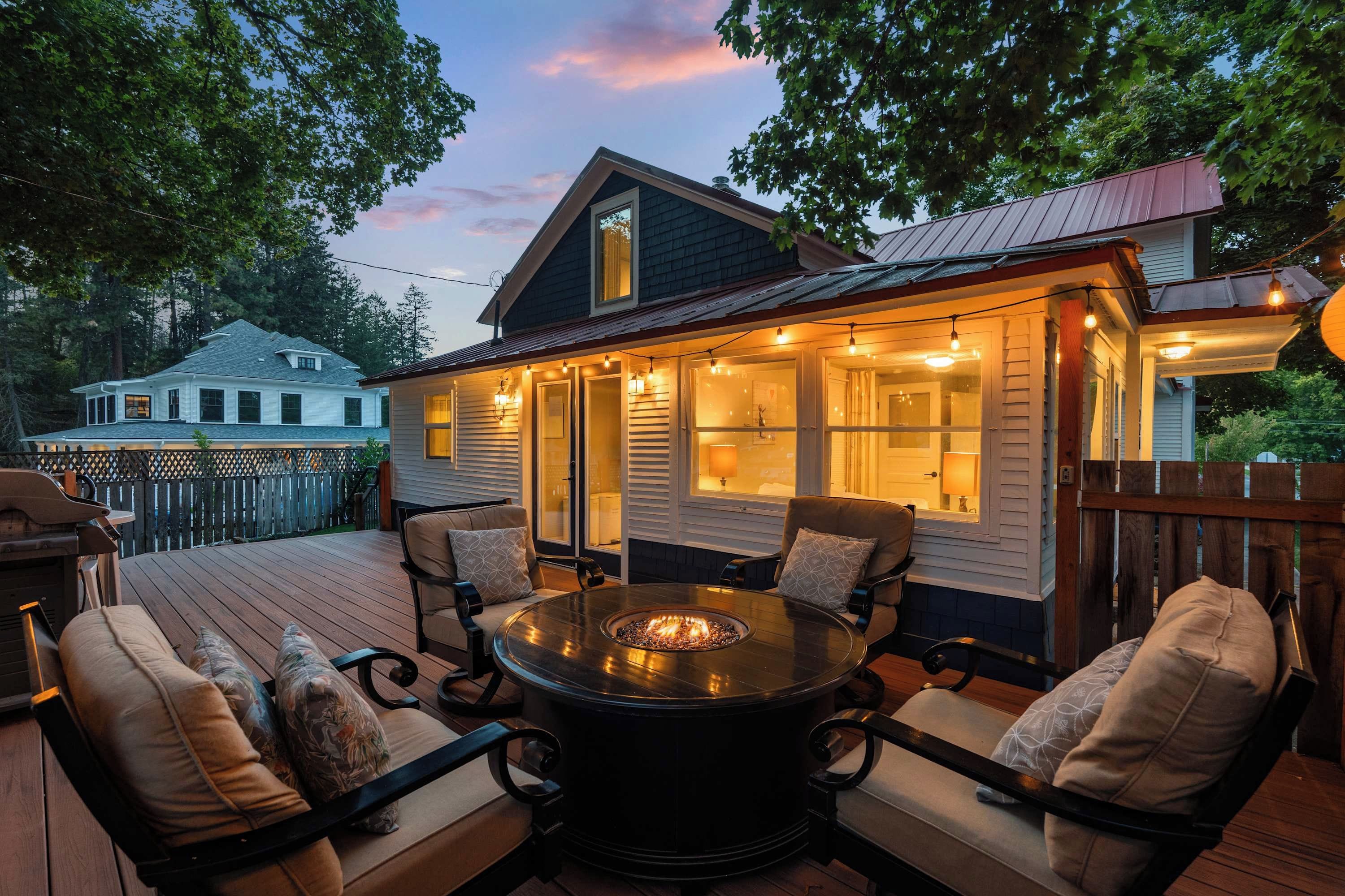 A deck with a round table and chairs, a fire pit, and a house with a porch lit by string lights.