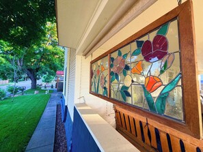 Porch detail: vivid stained-glass floral panel on the hanging swing, overlooking the tree-lined front yard - Classic CDA Charm with Porch | Walk to Downtown (Coeur d'Alene)