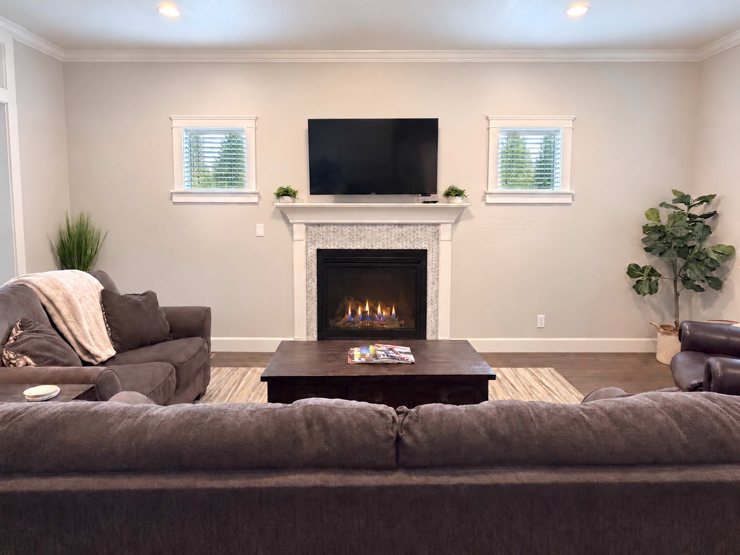 A living room with a fireplace, a television mounted above it, and two windows with blinds.