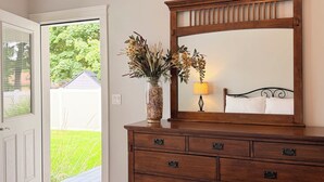 Un dormitorio con un aparador de madera, un espejo, un jarrón con flores secas y una lámpara de noche.