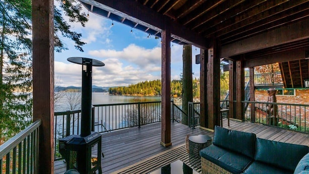 A covered deck with a view of a lake, wooden furniture, and a fire pit. - Lakefront Luxury | Dock, Hot Tub & Entertainment (Hayden Lake)