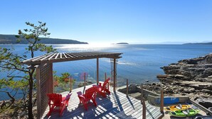 A wooden deck with red chairs overlooking a calm body of water, a rocky shoreline, and a clear sky. - Waterfront Serenity on the Sunshine Coast BC*
*90 day min rent due to BC Law (Halfmoon Bay)