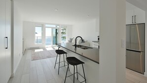 A modern kitchen with a central island, two bar stools, a stainless steel refrigerator, and a view of the outdoors through large windows. - Waterfront Serenity on the Sunshine Coast BC*
*90 day min rent due to BC Law (Halfmoon Bay)