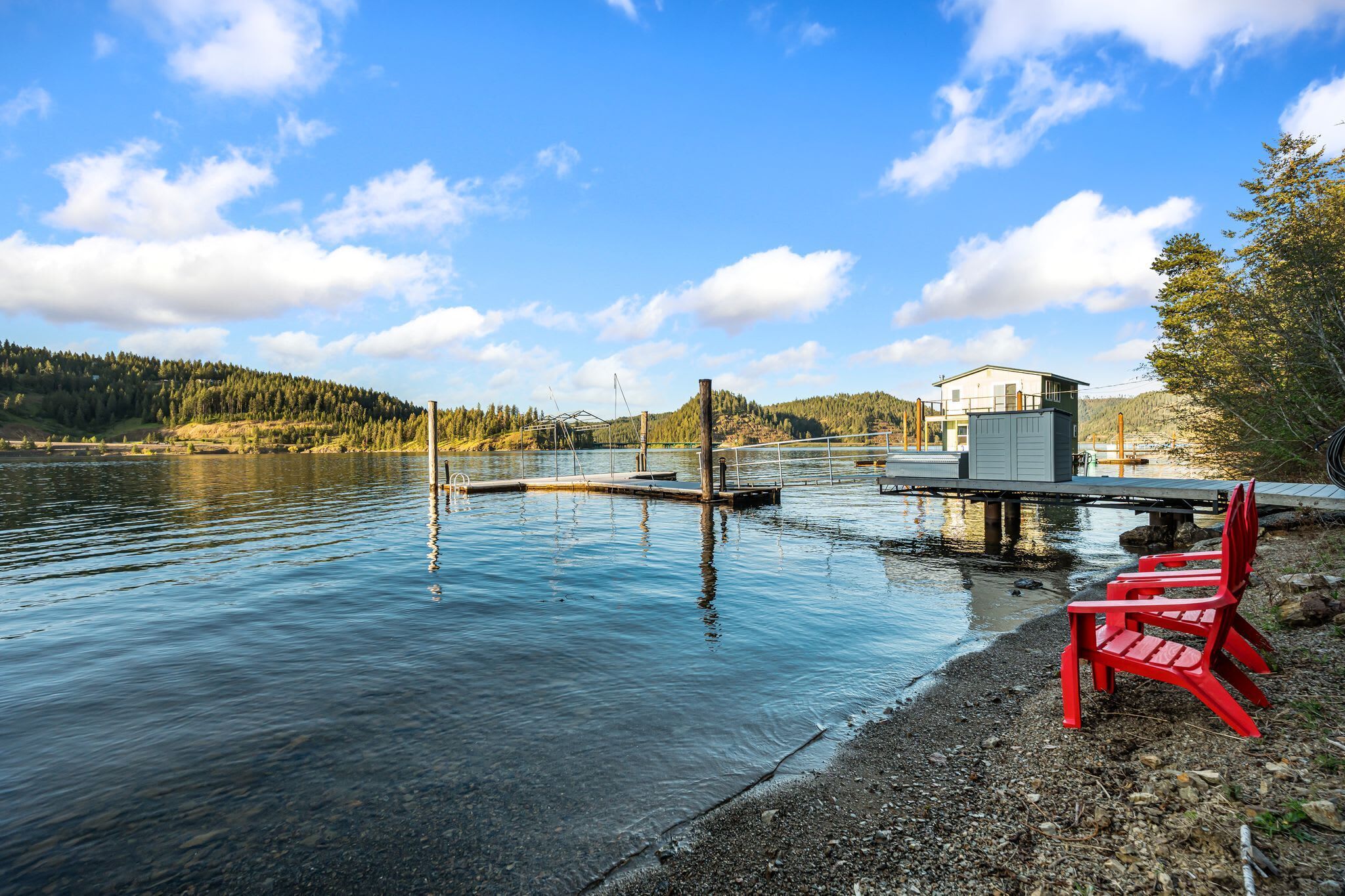 A lakeside view with a dock, a small house, and red chairs on the shore.