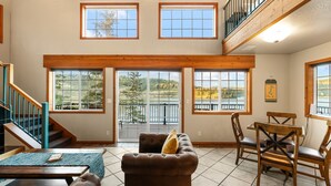 A spacious living area with a high ceiling, large windows, a staircase, and a dining area.