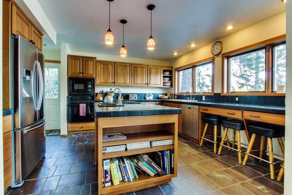 Una cocina moderna con armarios de madera, un refrigerador de acero inoxidable, un microondas y un horno empotrado. Hay tres lámparas colgantes, un reloj y una ventana con vistas a árboles.