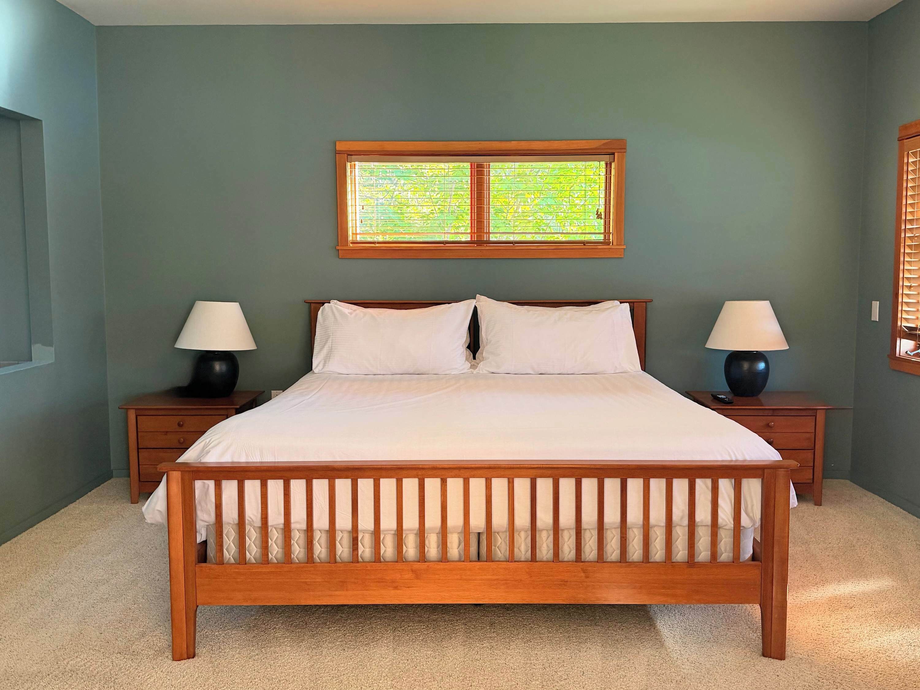 A bedroom with a wooden bed, two bedside tables with lamps, a window with blinds, and a carpeted floor.