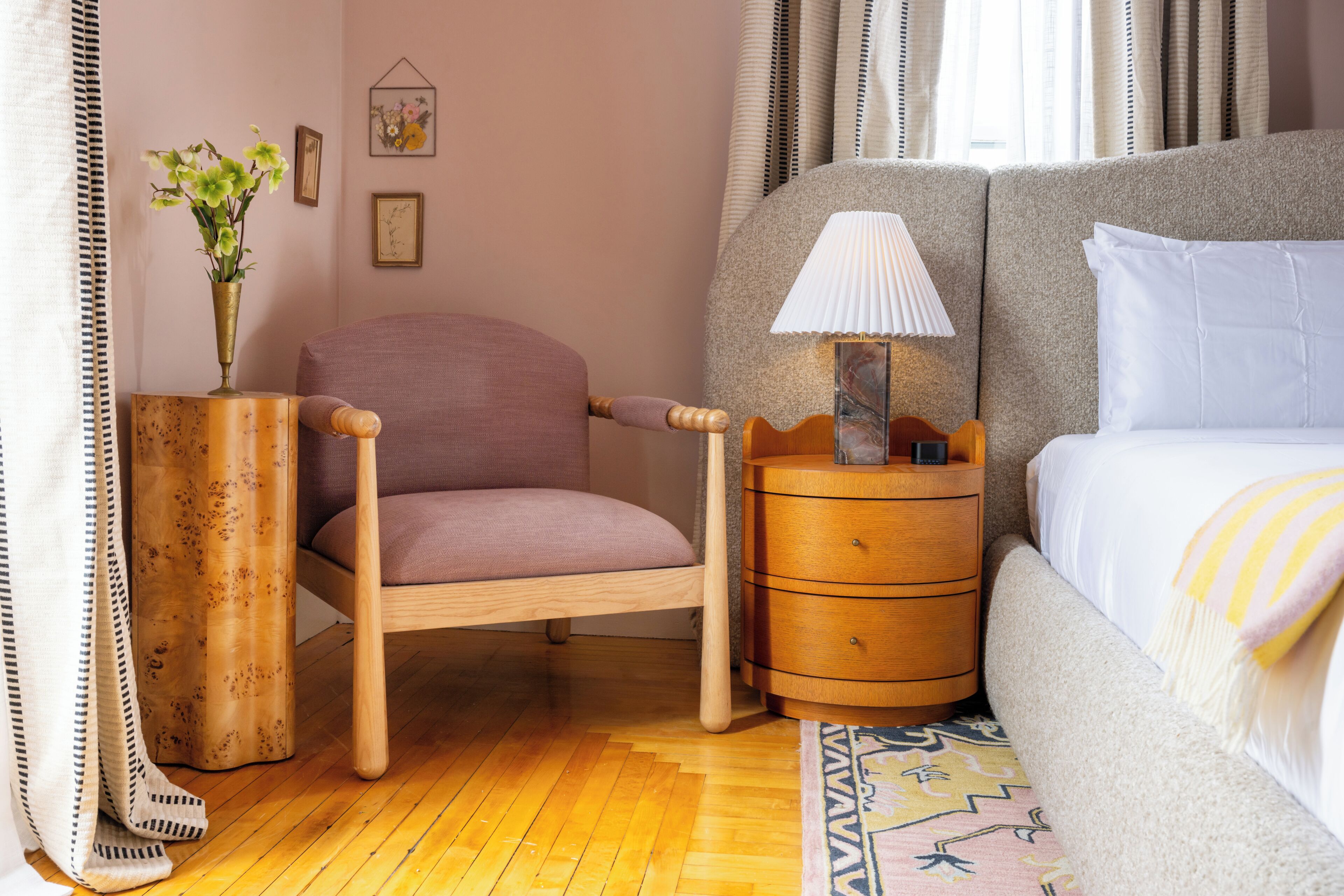 A bedroom with a bed, a chair, a nightstand, a lamp, a vase with flowers, and a wall with framed pictures.