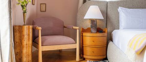 A bedroom with a bed, a chair, a nightstand, a lamp, a vase with flowers, and a wall with framed pictures.