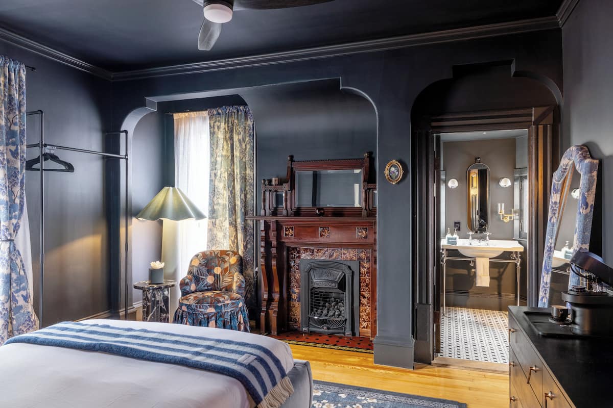 A bedroom with a bed, a chair, a fireplace, a mirror, and a bathroom visible through an archway.