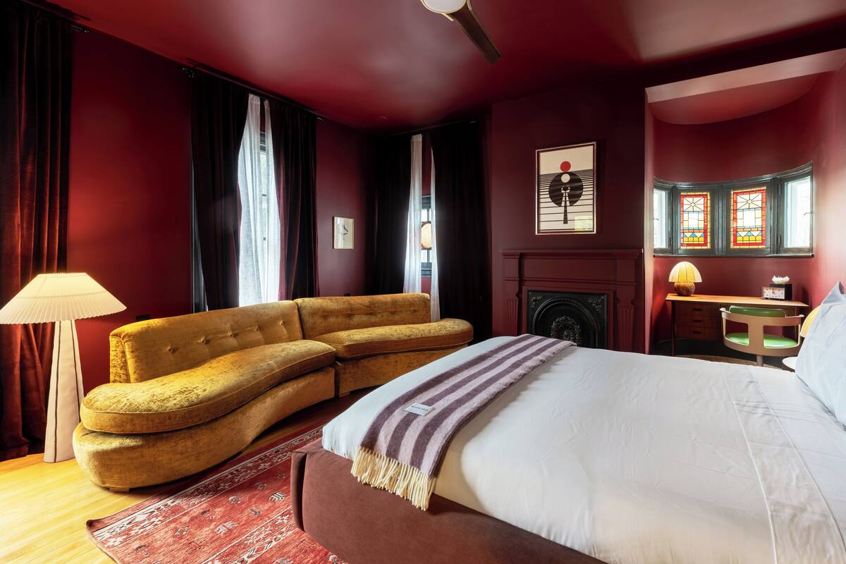 A room with a bed, a sofa, a lamp, a fireplace, and a desk.