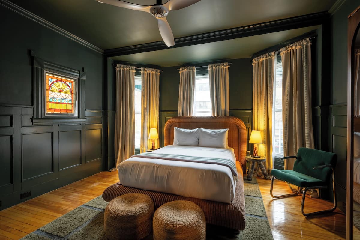 A bedroom with a large bed, a ceiling fan, a green chair, and a stained glass window.