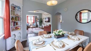 A dining area with a white table set for four, a bookshelf, and a television in the background. - Modern Midtown Getaway! Central location for 12! (Omaha)