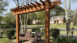 A wooden pergola with a bench underneath, surrounded by greenery and a brick pathway.