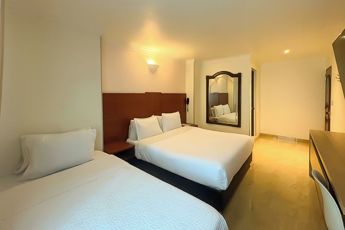 Habitación de hotel con dos camas, cabecero de madera, mesita de noche, espejo y lámpara de pared.