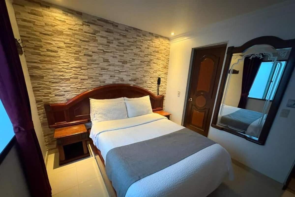 A hotel room with a stone wall, a wooden bed, a nightstand, a mirror, and a door.