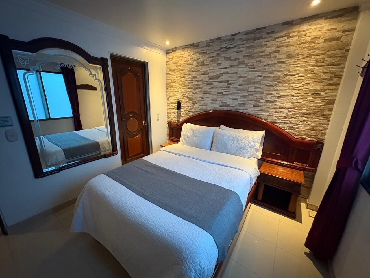 Una habitación de hotel con una cama, cabecero de madera, pared de piedra, espejo y mesita de noche.