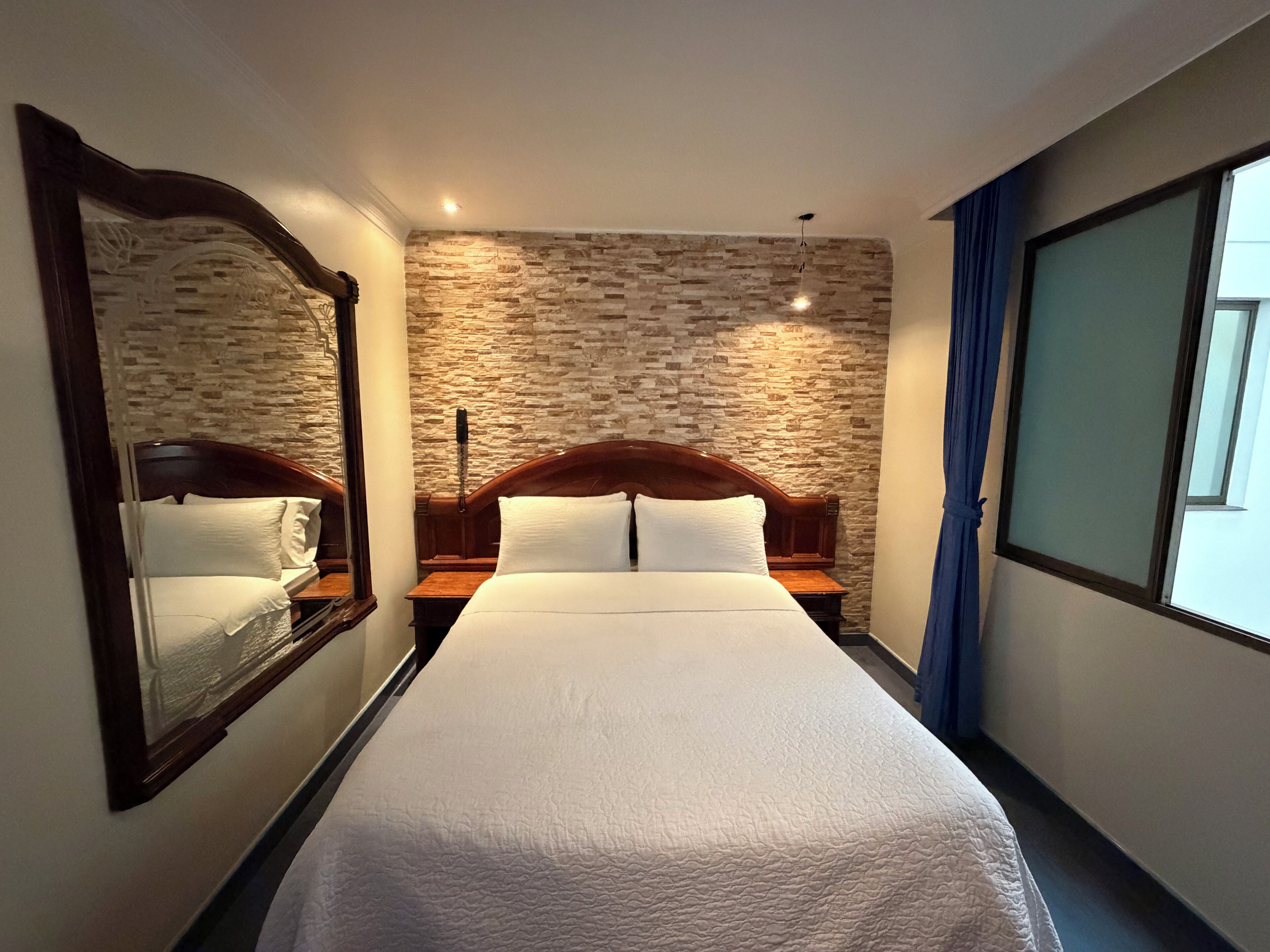 Ein Hotelzimmer mit einem großen Bett, einer Steinswand, einem Nachttisch, einem Spiegel und einem Fenster mit Vorhängen.