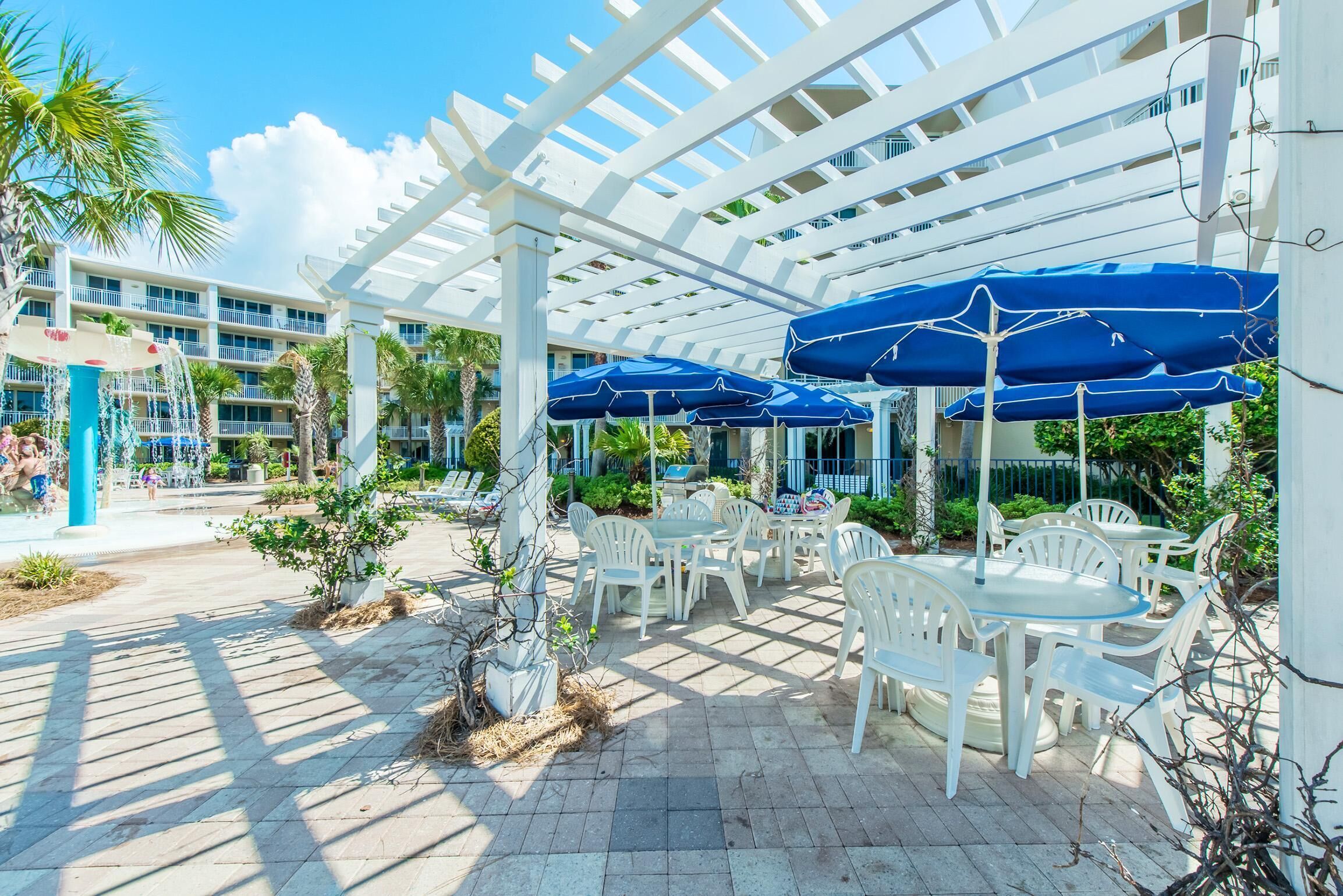 An outdoor patio with white chairs and blue umbrellas, surrounded by a white pergola structure.