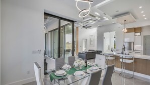 Et moderne spiseområde med et glasbord, hvide stole og en grøn løber. Der er et køkkenområde med en bar og en loftsvifte.