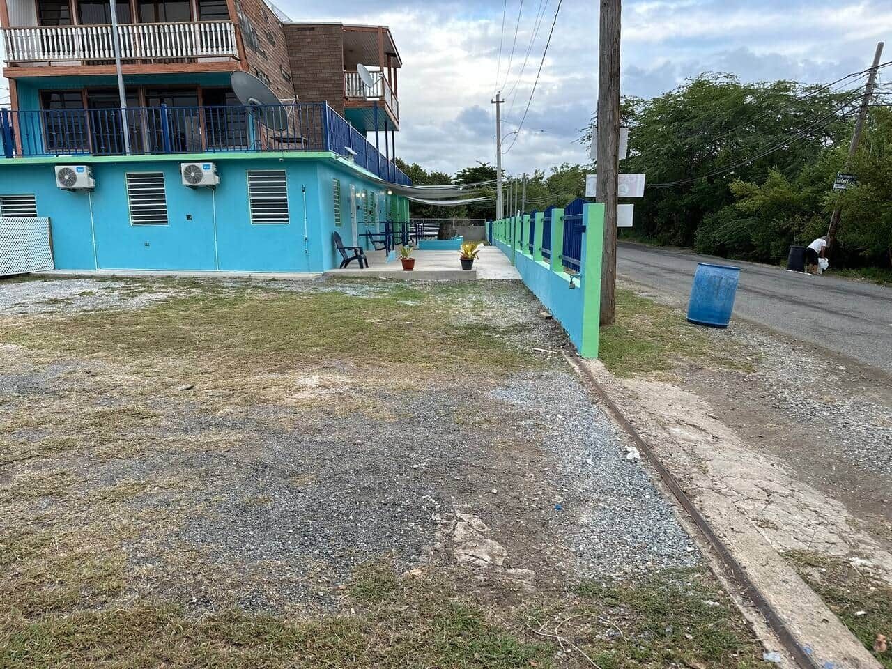 Edifici amb façana blava, un balcó i una zona pavimentada amb un camí que condueix a una carretera.