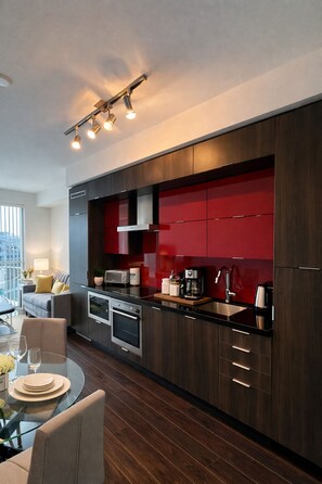 Una cocina moderna con armarios de madera oscura, una pared de acento roja y electrodomésticos de acero inoxidable. Hay un comedor con una mesa y sillas de vidrio.