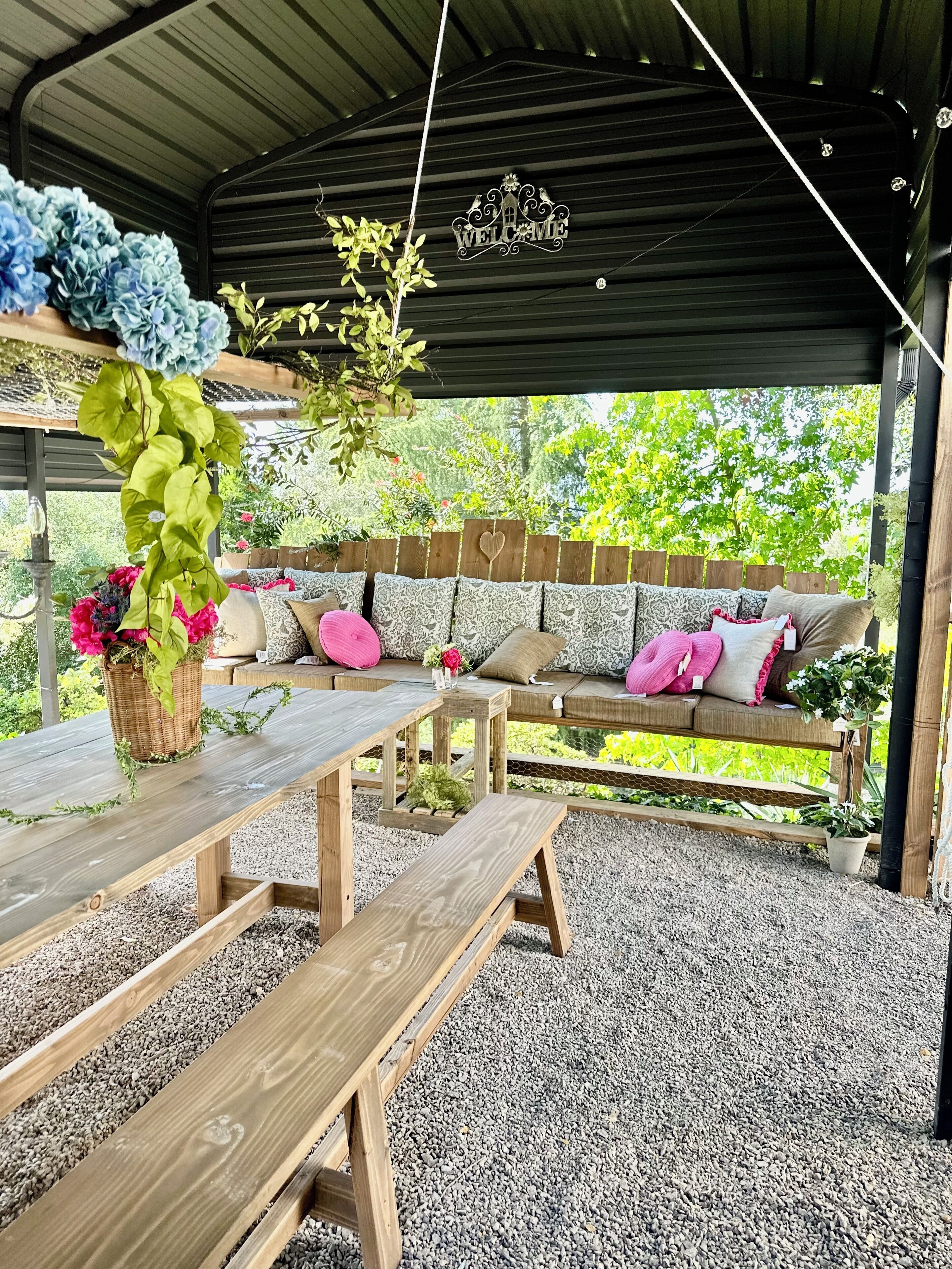 An outdoor covered seating area with a wooden bench, a cushioned sofa with patterned cushions, and a wicker basket with flowers.