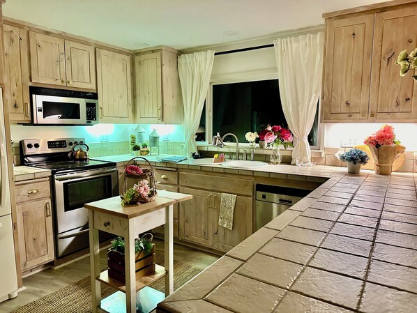Una cocina con armarios de madera, microondas, estufa, fregadero y una mesita con plantas en macetas.