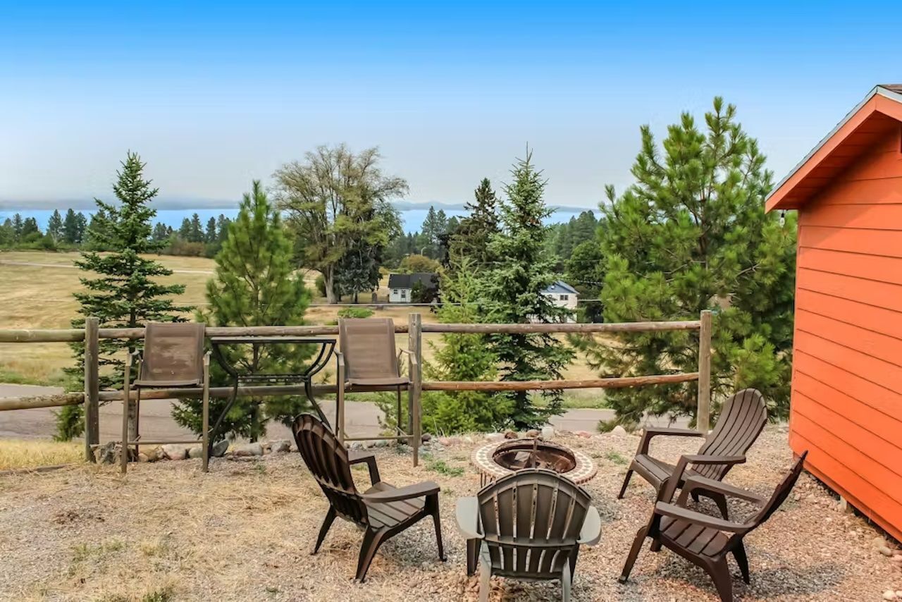 An outdoor seating area with Adirondack chairs and a fire pit, overlooking a scenic landscape with trees and a distant view of mountains.