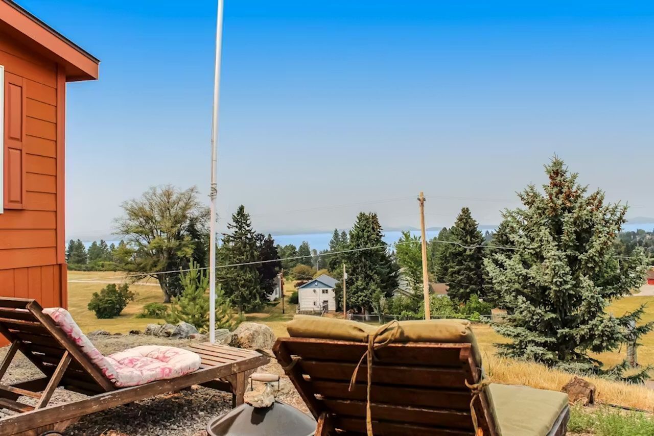 A wooden deck with lounge chairs and a table, overlooking a rural landscape with trees and a small house in the distance.