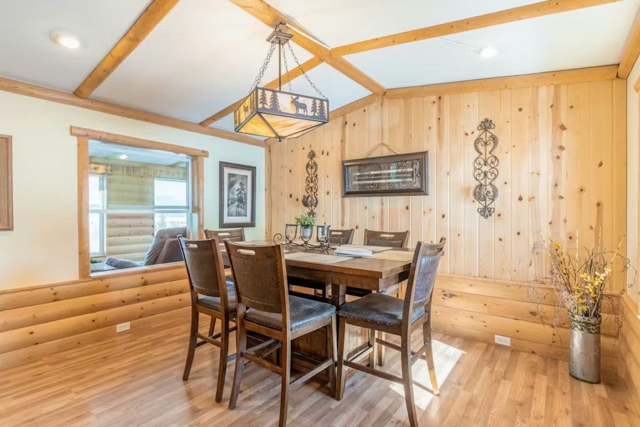 A wooden-paneled dining room with a large table and chairs, a hanging light fixture, and framed pictures on the walls.